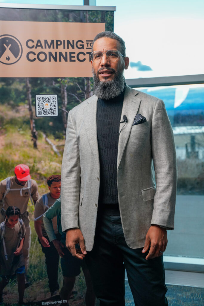 Man with gray beard and glasses in a light gray blazer speaking at a 'Camping to Connect' event, outdoors with a banner in the background.